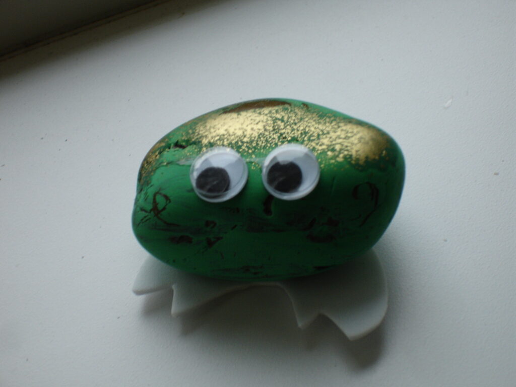 A rock has been painted green and gold, and googly eyes have been attached to it and underneath, felt feet.