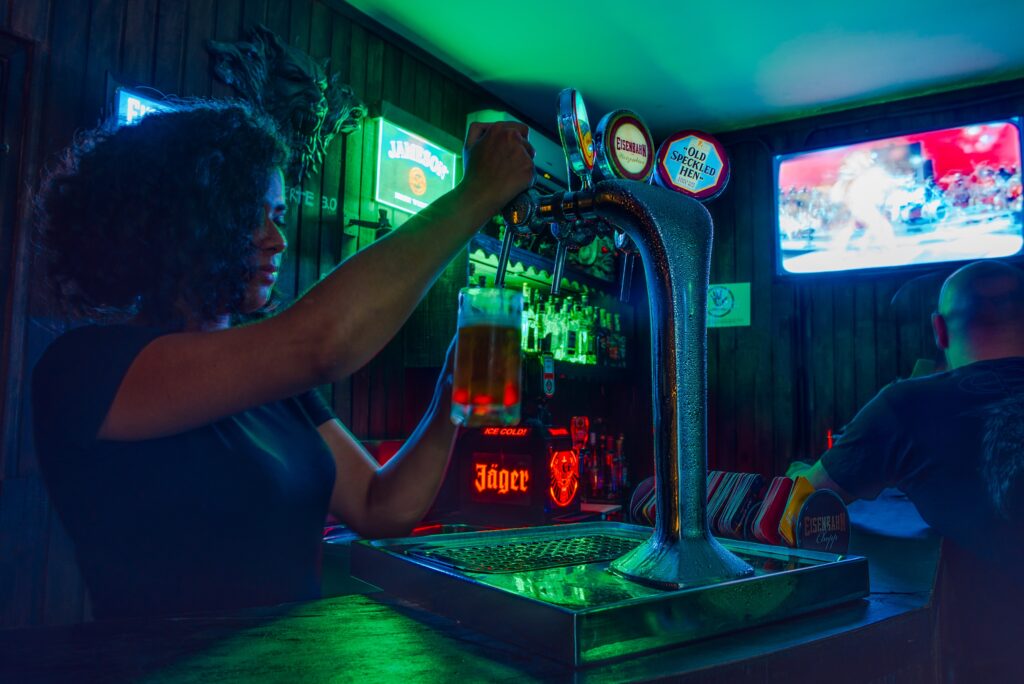 A person with curly hair pulls a beer tap to fill a pint glass behind a bar counter.