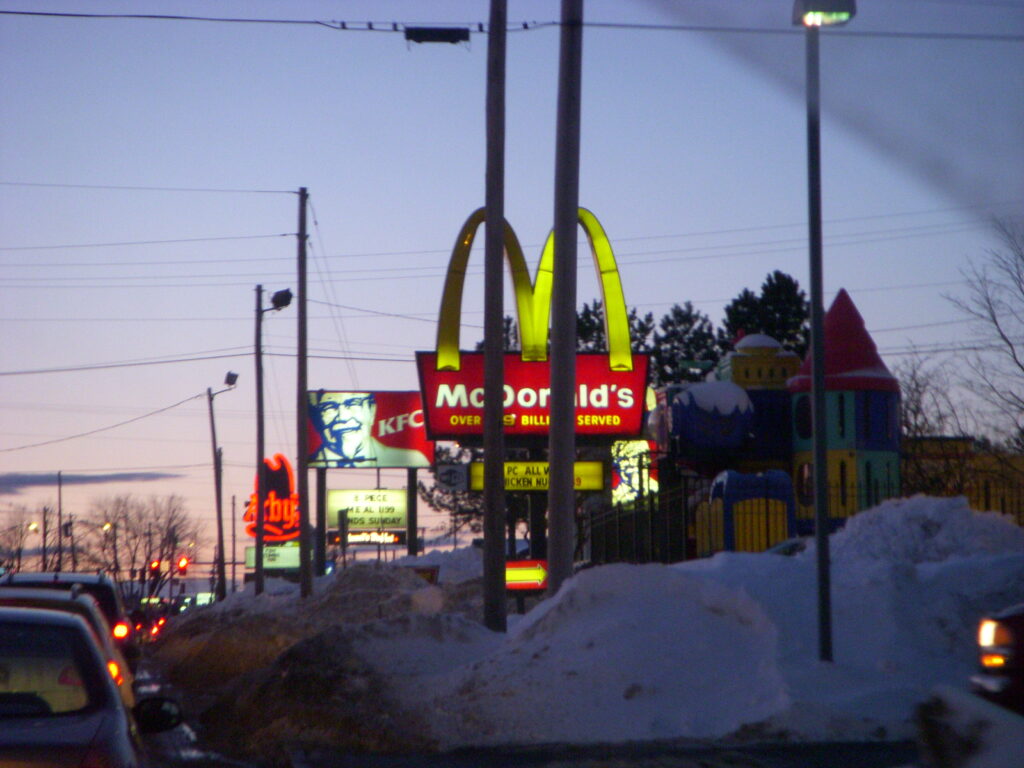 A photograph features a roadside lined with the lit promotional signs of McDonalds, KFC, and Arby’s, one after the other.