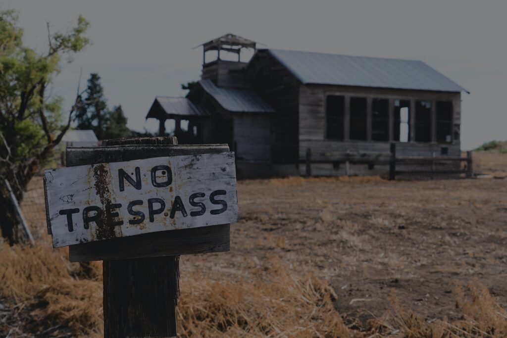 A white wooden sign reads “No Trespass” in front of an old gray wooden structure on a dirt plot sprinkled with yellowed grass.