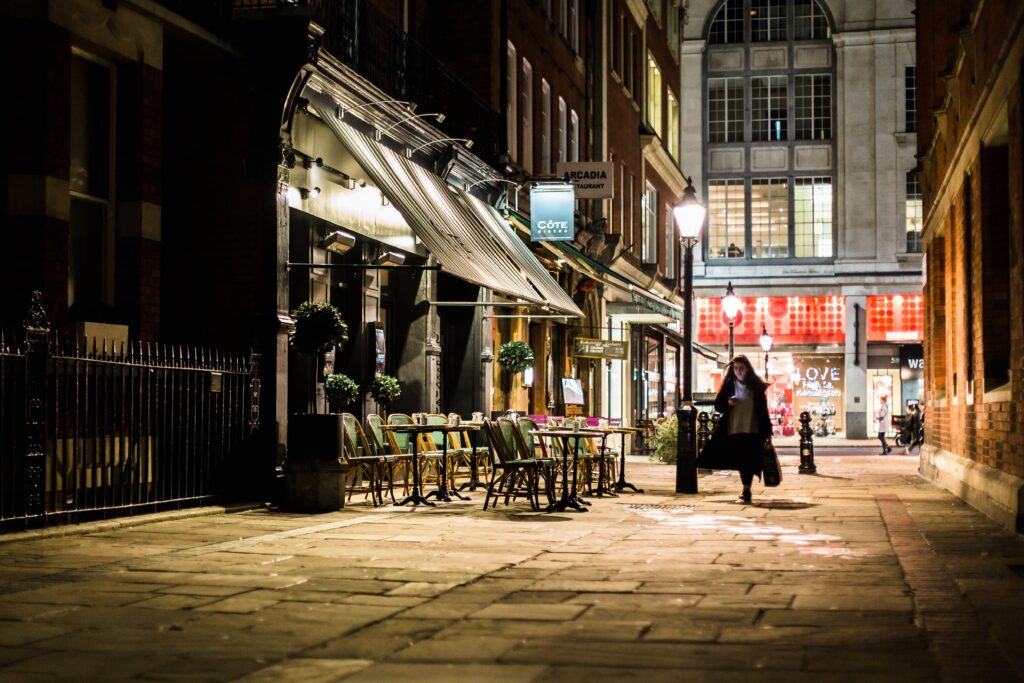 A photograph features a person in a peacoat carrying a bag and walking down a side street past a closed café at night.
