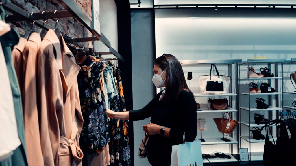 A person wears a white face mask and looks through a rack of clothing in a store. A shelf of shoes is in the background.