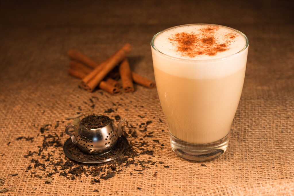 A small, clear glass holds light-brown liquid with white foam and brown powder on top. A pile of cinnamon sticks sits beside the glass.