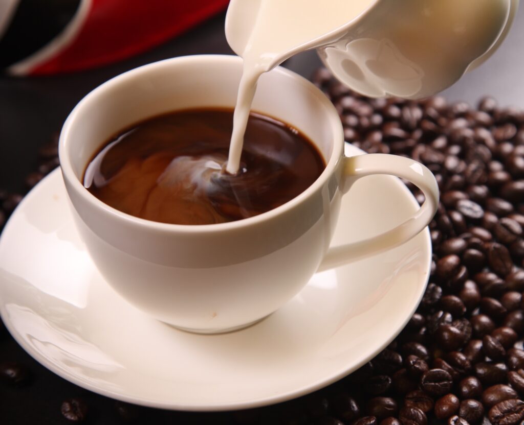 : A close-up photograph features a small white cup with dark brown liquid on a white saucer with white liquid being poured into it from a small pitcher.