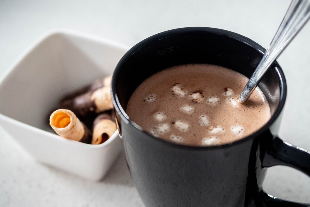 A black mug with brown liquid, floating marshmallows, and a metal spoon in it rests on a white surface. A small bowl of pastries sits beside it.