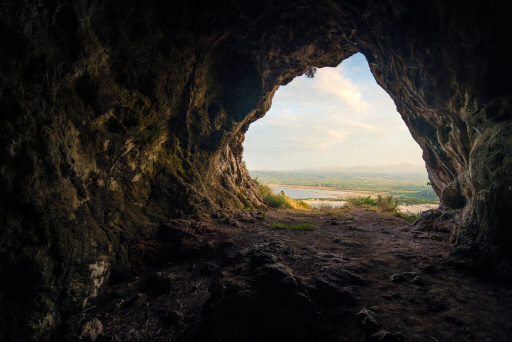 The view looks out of a dark cave onto blue water and a sandy beach.