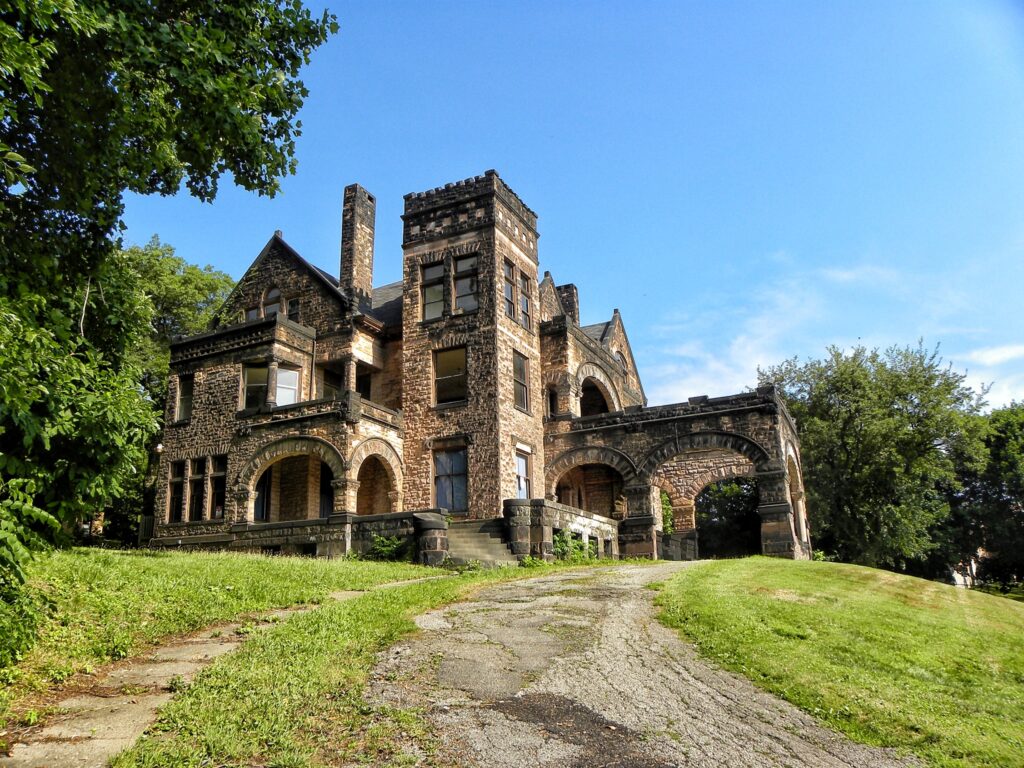 A large brown, brick mansion is shown with a grassy yard in front and a blue sky overhead.