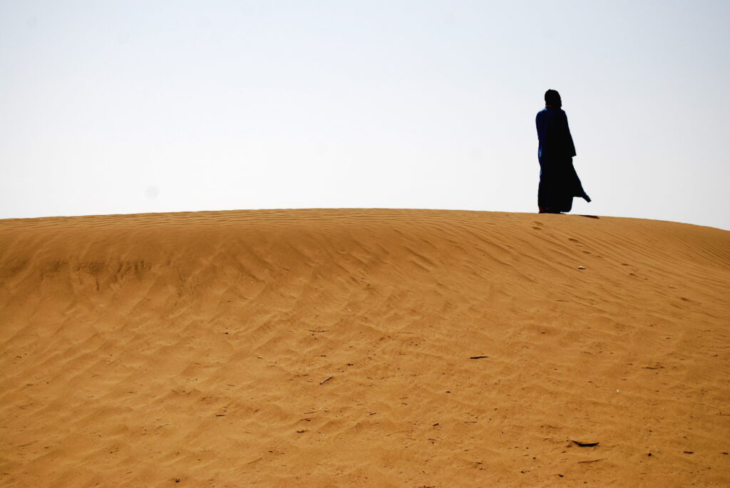 A person dressed in a black robe stands on a sand dune.