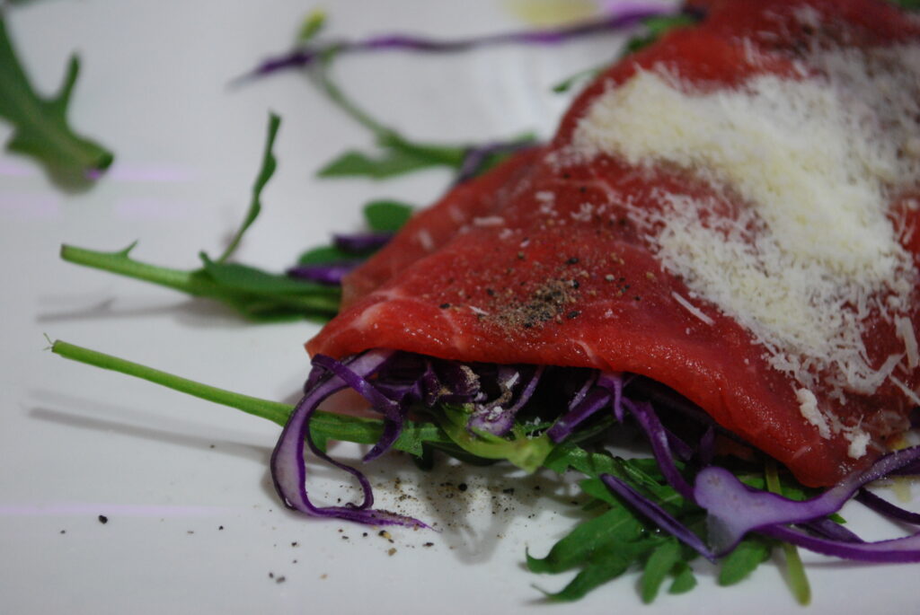 A thin slice of raw meat and green and purple garnish are shown on a white plate.