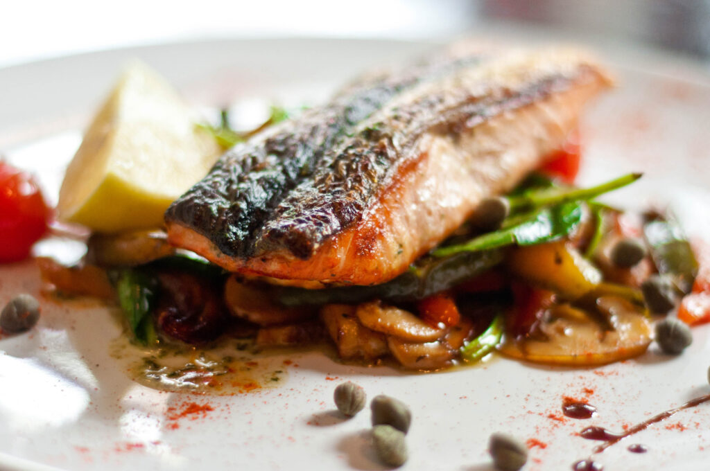 A piece of fish laying on a bed of cooked vegetables, with a sliced lemon on the side.