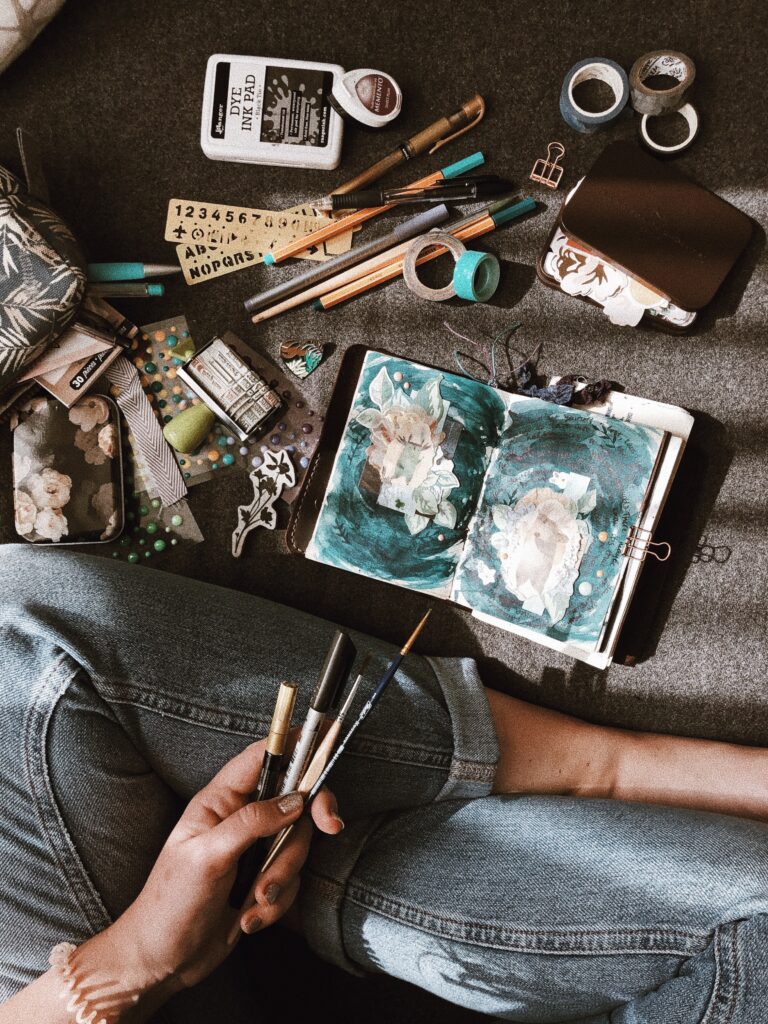From above, a photograph features a journal with its open pages painted with brushes and paints to the right and a person, sitting cross-legged, holding more brushes to the left.