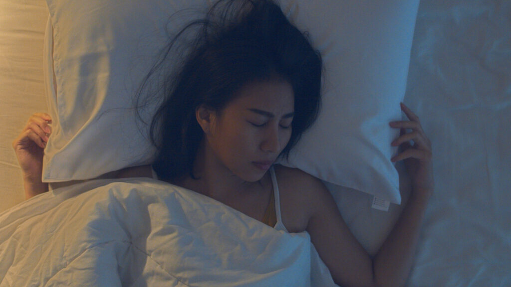 A person with long, black hair sleeps on white sheets with matching pillow and blanket.