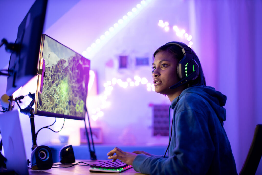 A person with straight, shoulder-length hair wears headphones in a bright purple room and stares at two desktop computer screens while typing on a keyboard.