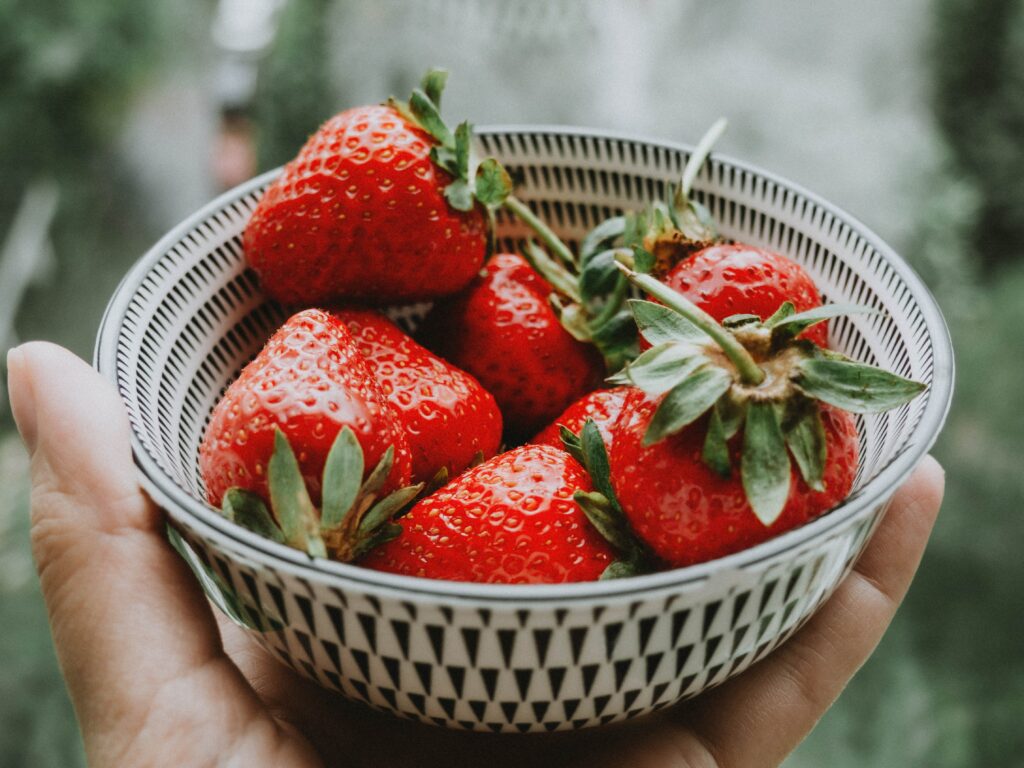 A close-up shows a small white-and-green bowl full of strawberries held up by a person’s hand.