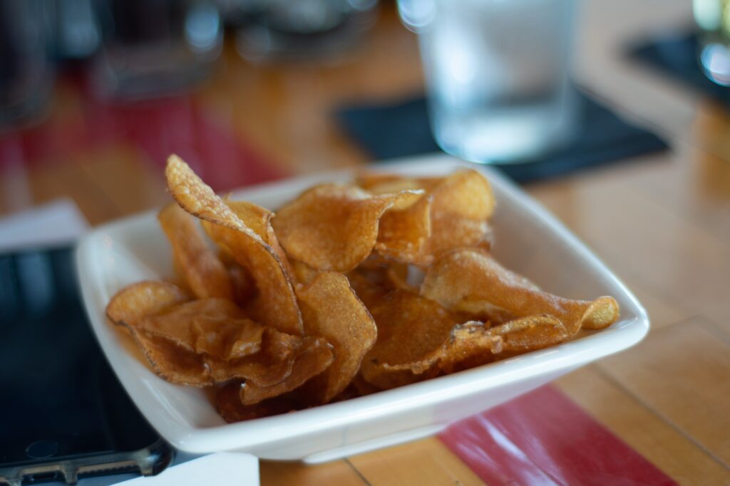 A small white bowl holds potato chips on top of a wooden table.
