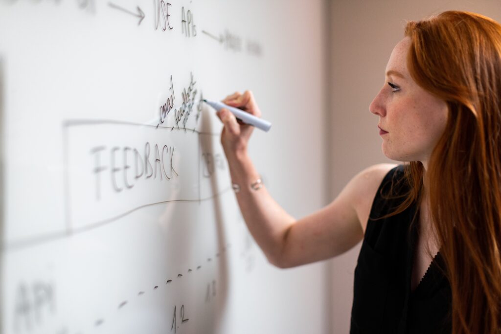 A person with long, red hair writes on a whiteboard with a marker.