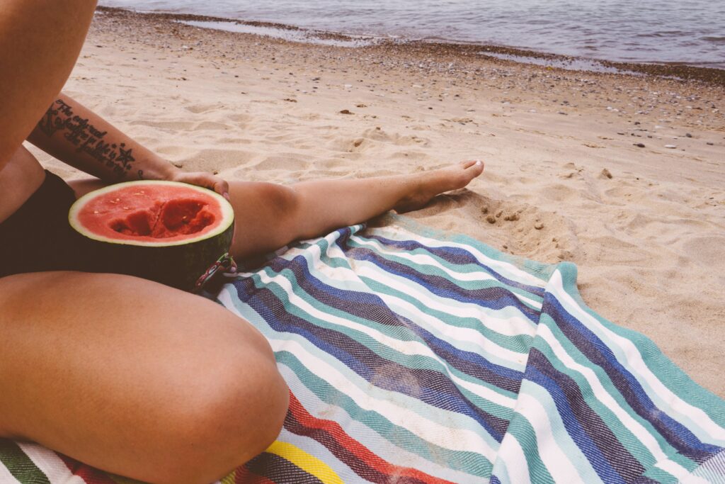 A watermelon half nests in the lap of a person sitting on a beach towel on a sandy shore, with water in the background.