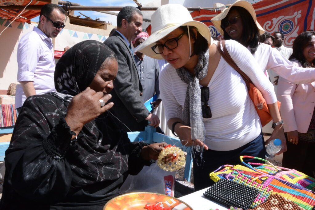 In a crowded marketplace, a person sits in front of a craft table. They pull thread tight with one hand from an object they hold in their other hand as they chat with a bystander.
