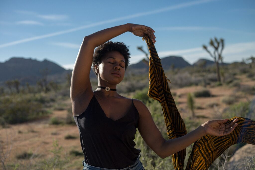 A person with short curly hair twirls a patterned scarf over their head against a background of sand, cacti, and a mountain in the distance.