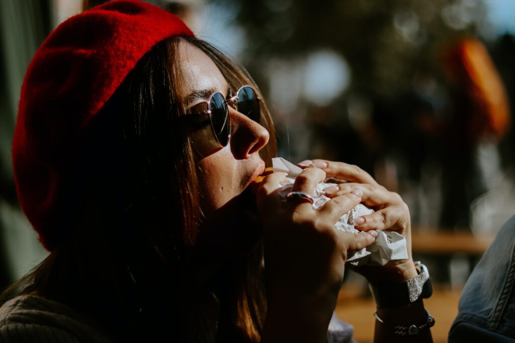 A close-up shows a person with long hair and a red beret taking a big bite of a sandwich.