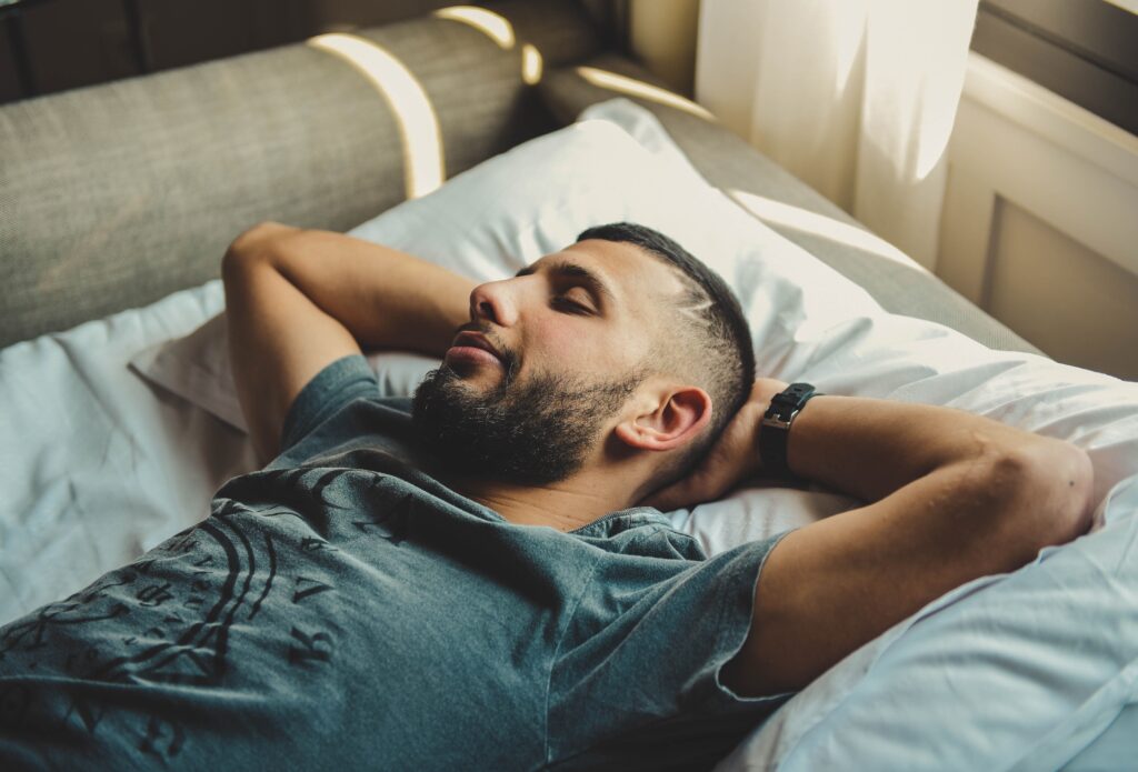 A photograph features a person with short hair, a mustache, and beard smiling while lying in bed with their eyes closed and their hands behind their head.