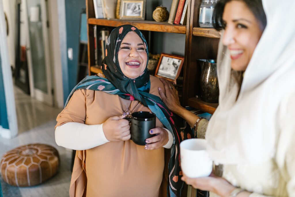 Two smiling people wearing headscarves hold mugs and face each other.