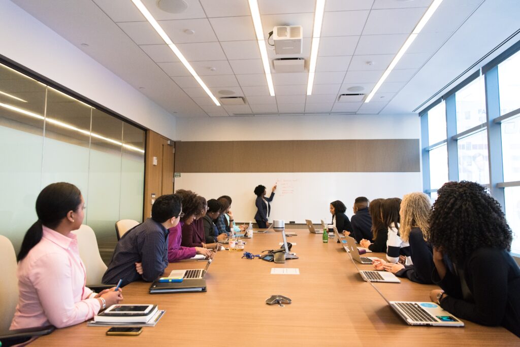 A long, wooden table is lined with people seated in front of laptops. One person stands at the table’s far end, pointing at a whiteboard on the wall.