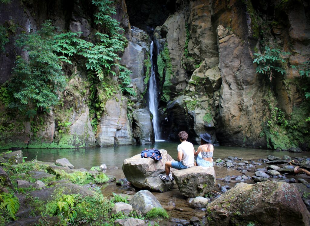 A photograph features two people sitting on a rocky cliff and looking over a lake at a waterfall on its other side.