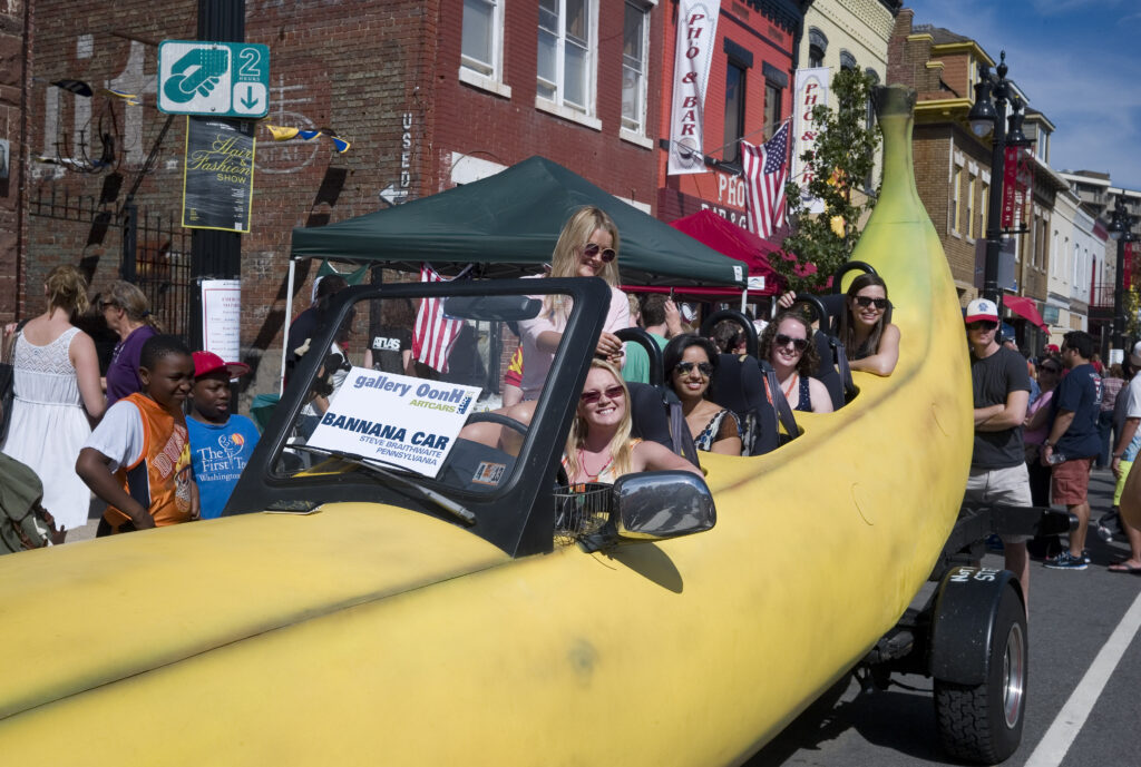 A photograph features several people smiling in a car shaped and painted yellow to resemble a banana with a sign on front that reads, “Banana Car.”
