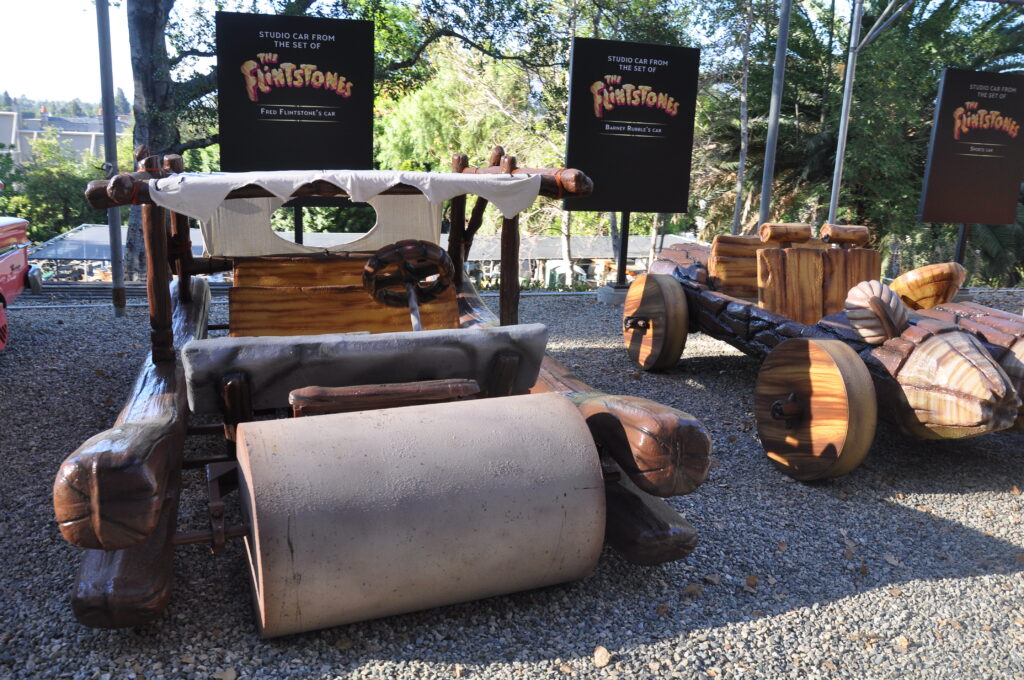 Two vehicles made of wood and a cloth roof sit next to one another. Each has one large cylindrical wheel at its front. They are in front of a sign that reads, “Studio Car from the set of The Flintstones.”