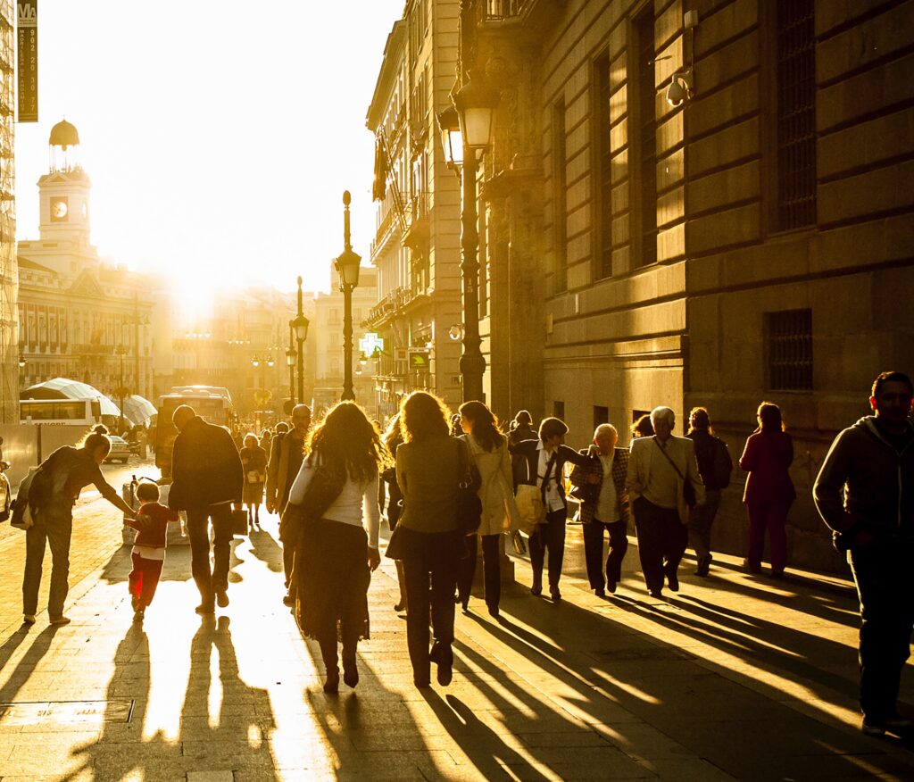 Many people walk down an urban street, with the sun setting in the background.