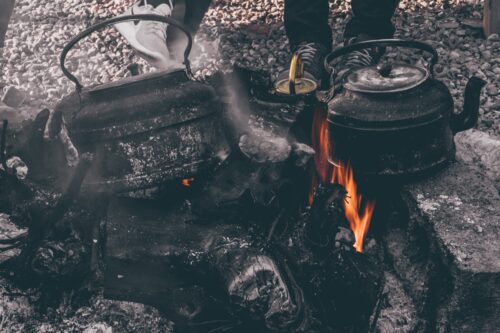 Surrounded by gray cobblestones, two black kettles sprinkled with ash sit over blackened logs and open orange flames. Two pairs of sneakers on people’s feet are visible just behind the fire close to another kettle.