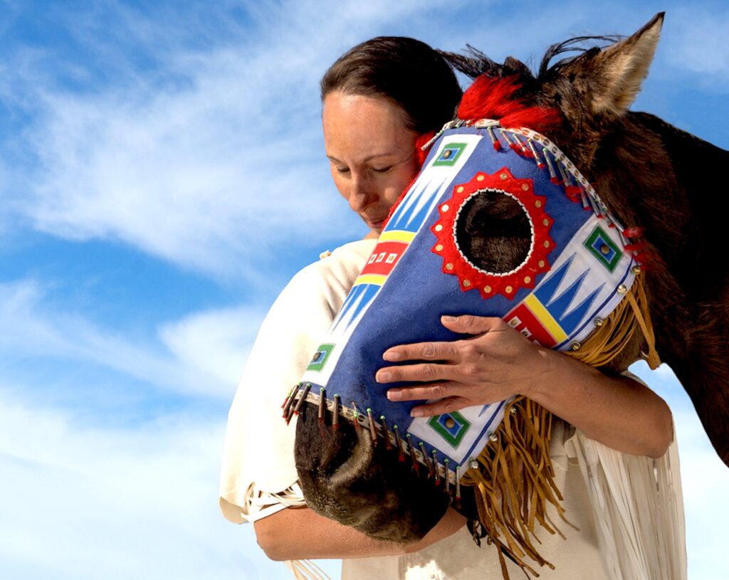 A person in a white poncho hugs the head of a horse wearing a colorful mask against a blue sky with white clouds.