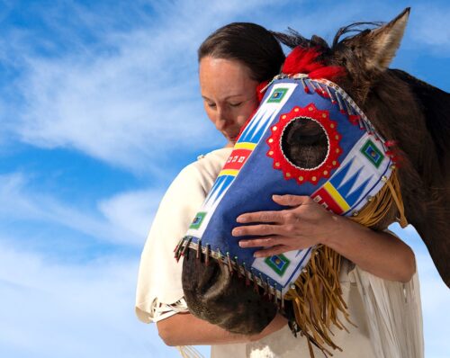 A person in a white poncho hugs the head of a horse wearing a colorful mask against a blue sky with white clouds.