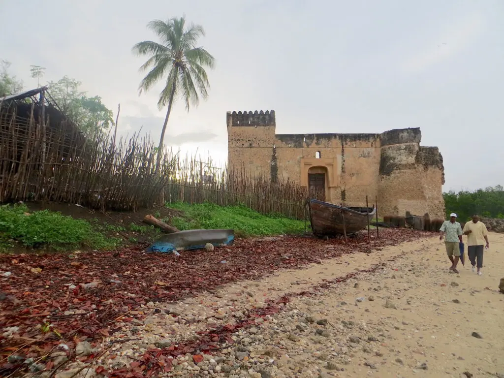 Two people walk on a sandy shore with a large stone castle and wooden gate to their right.