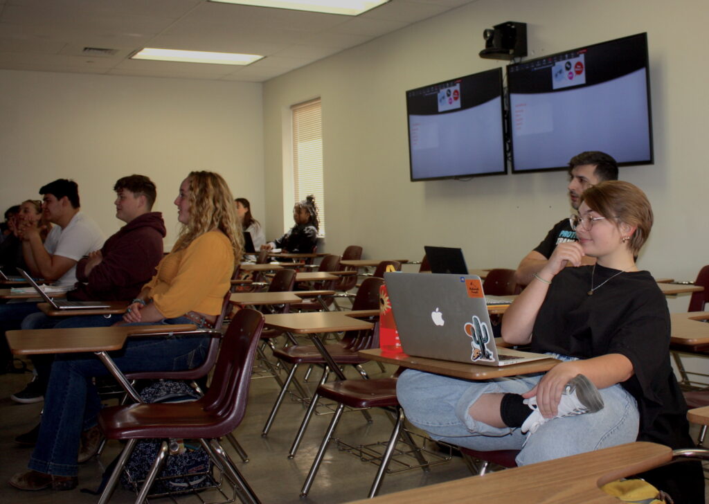 Several students looking toward the front of a classroom sit at wooden desks with open laptops on them. Two monitors hang on the back wall.