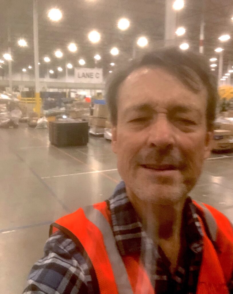 A person with short brown hair and facial wrinkles wearing a blue plaid collared shirt and bright orange vest stands in a warehouse.