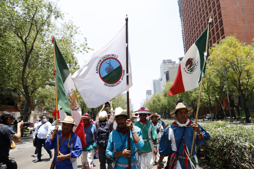 Several people wearing colorful clothing and brimmed hats march in a group behind three who are displaying white, green, and red flags emblazoned with logos and the word “Wixárika.”