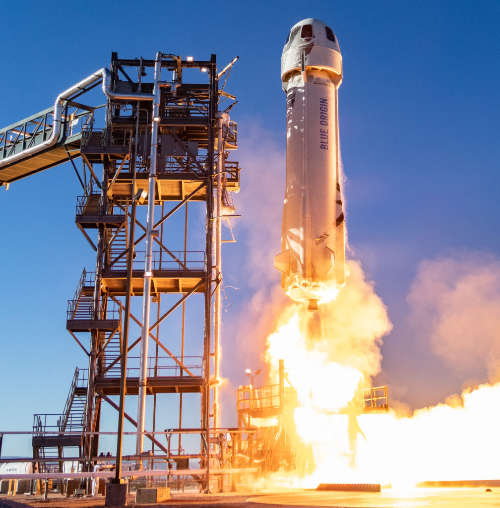 Flames and smoke emerge from a white long, domed rocket leaving a launchpad on a large cement plot. A large metal structure lies on its left.