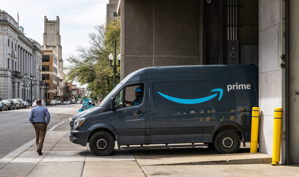 A person on a sidewalk passes a large blue van with a blue arrow and the word “prime” on its side driven by a person wearing a blue hat. The van’s rear is backed into an open garage.