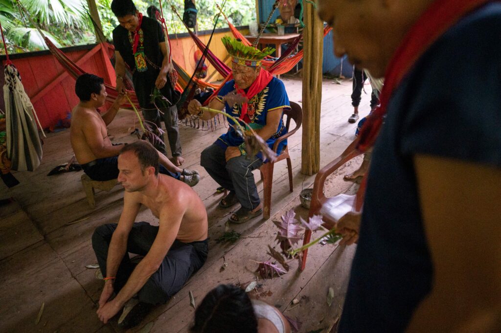 In a semi-open space in the Amazon, an elder wearing a headdress waves a long plant next to a shirtless person sitting on the ground. Several other people interact around them.
