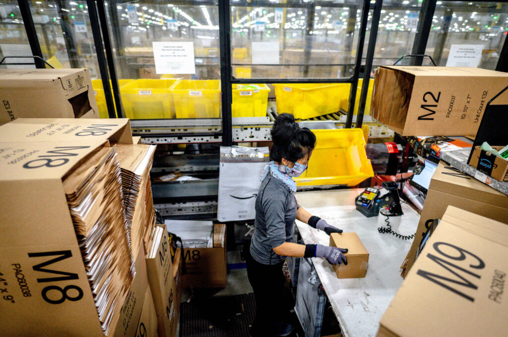 In a warehouse, a person with a black ponytail, face mask, and gloves assembles a cardboard box on a workstation amid neat piles of unfolded cardboard boxes.