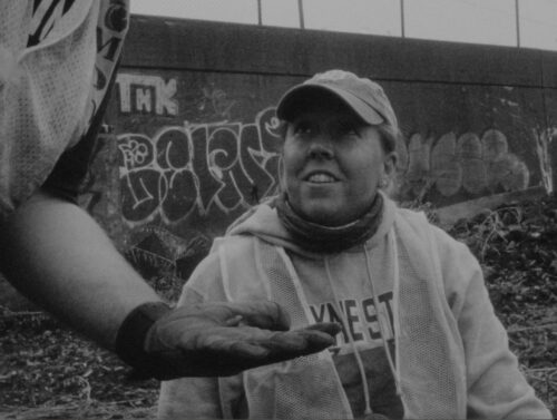 A black-and-white film still features a person wearing a hoodie, clear vest, and cap looks upward at the face of a person holding small objects in front of them with a gloved hand. The two are on rocky terrain in front of a graffiti-covered stone wall.