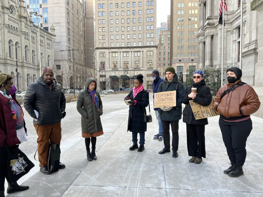 A group of people in jackets and hats gather in an arc on a sidewalk in front of tall concrete buildings. Two hold carboard signs. One reads “Community Control” and the other says “Return the Remains.”