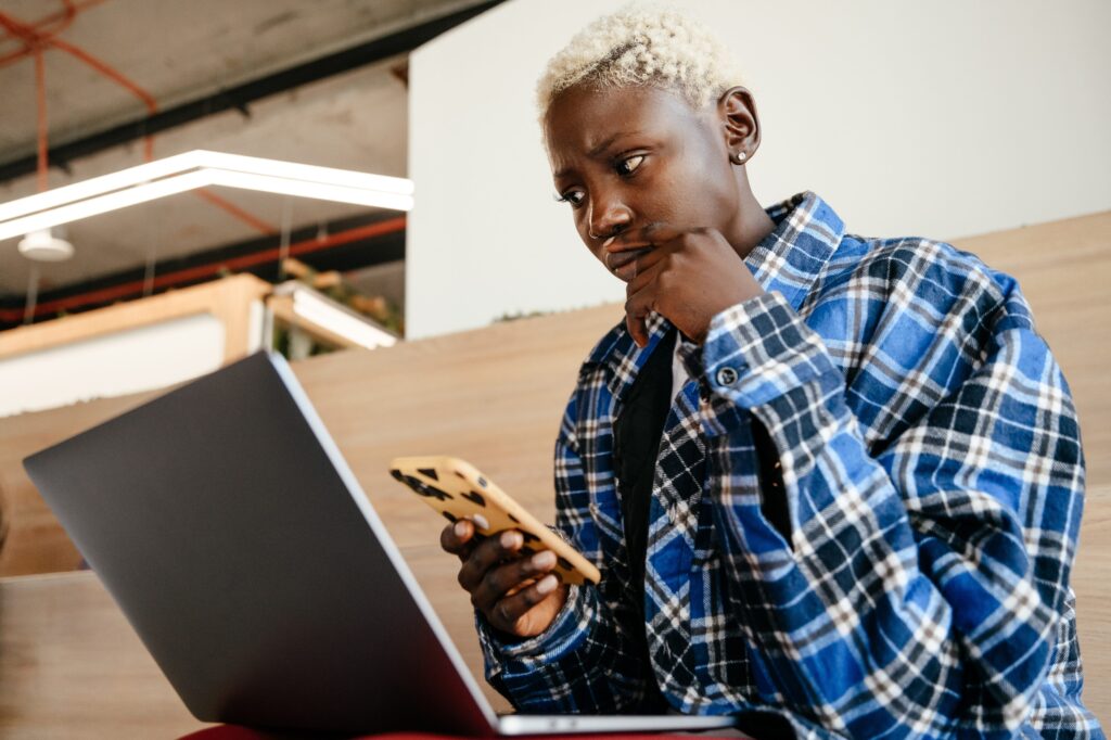 A young adult with short, platinum-blonde hair wearing a blue-and-white plaid flannel shirt sits on a long wooden bench in front of an open silver laptop. They look at the screen puzzlingly while holding up a yellow cellphone.