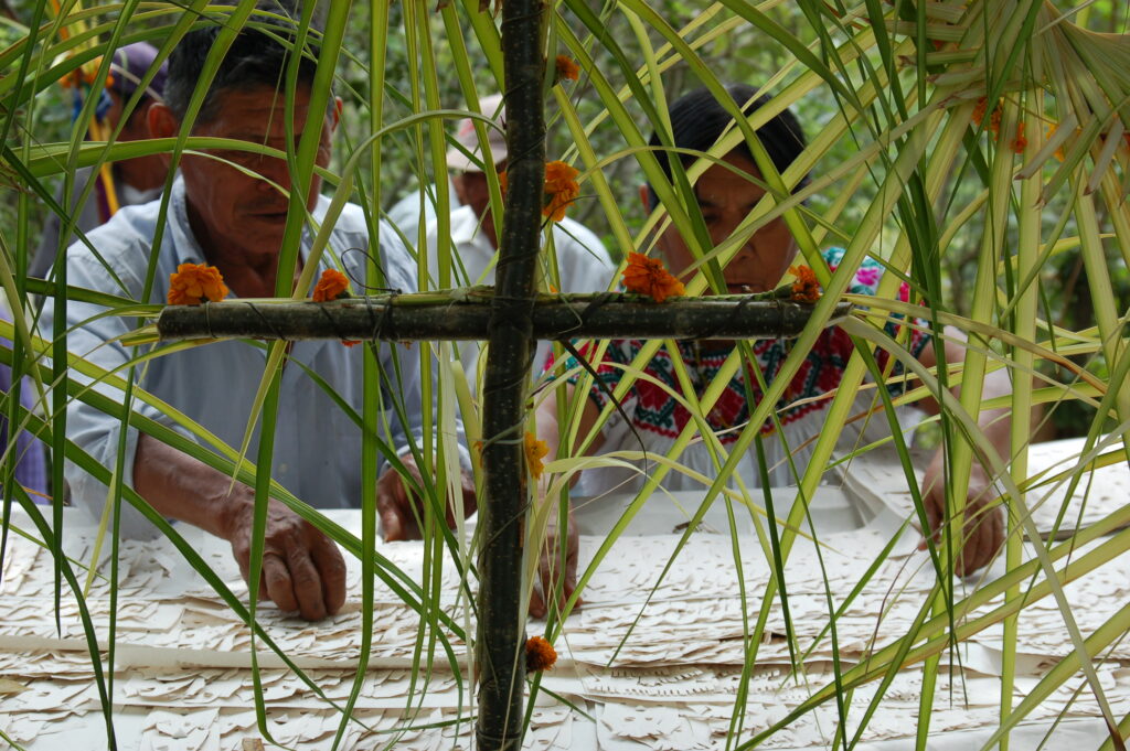 In the photo’s foreground, a cross made of sticks surrounded by palm leaves rises next to a table that holds a large pile of small figures cut from white paper. People organize the paper in the background.