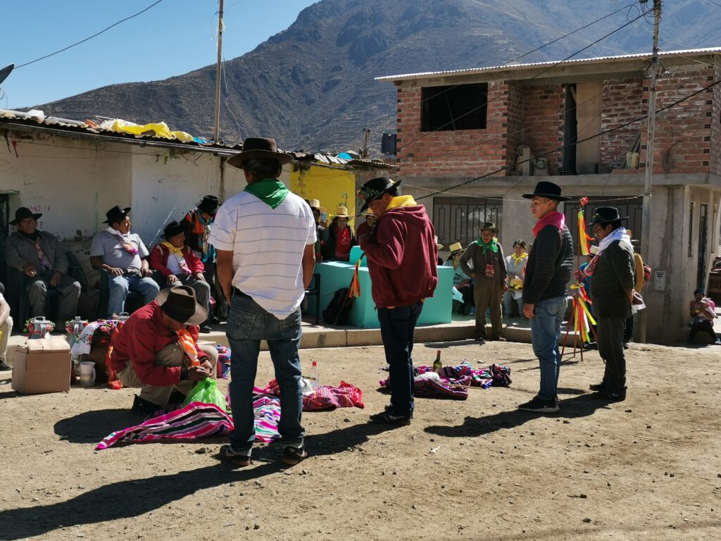 Personas sentadas alrededor de cuatro personas que llevan sombreros de alas anchas paradas en una plaza. Al frente de las cuatro personas hay una persona agachada y tocando un objeto verde que está encima de una tela con diseños.