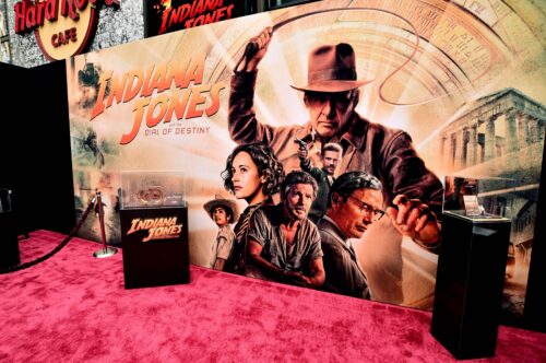 Behind a red carpet, a large poster features a person in a brown brimmed hat holding up a whip over a cluster of other people. A pillared building lies in the background opposite yellow and orange text that reads, “Indiana Jones and the Dial of Destiny. It is flanked by display cases, one holding a bronze time dial.