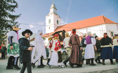 A crowd of people wearing head coverings and long dresses or robes outfits decorated with colorful accessories walk in line through public streets past a white steepled building.