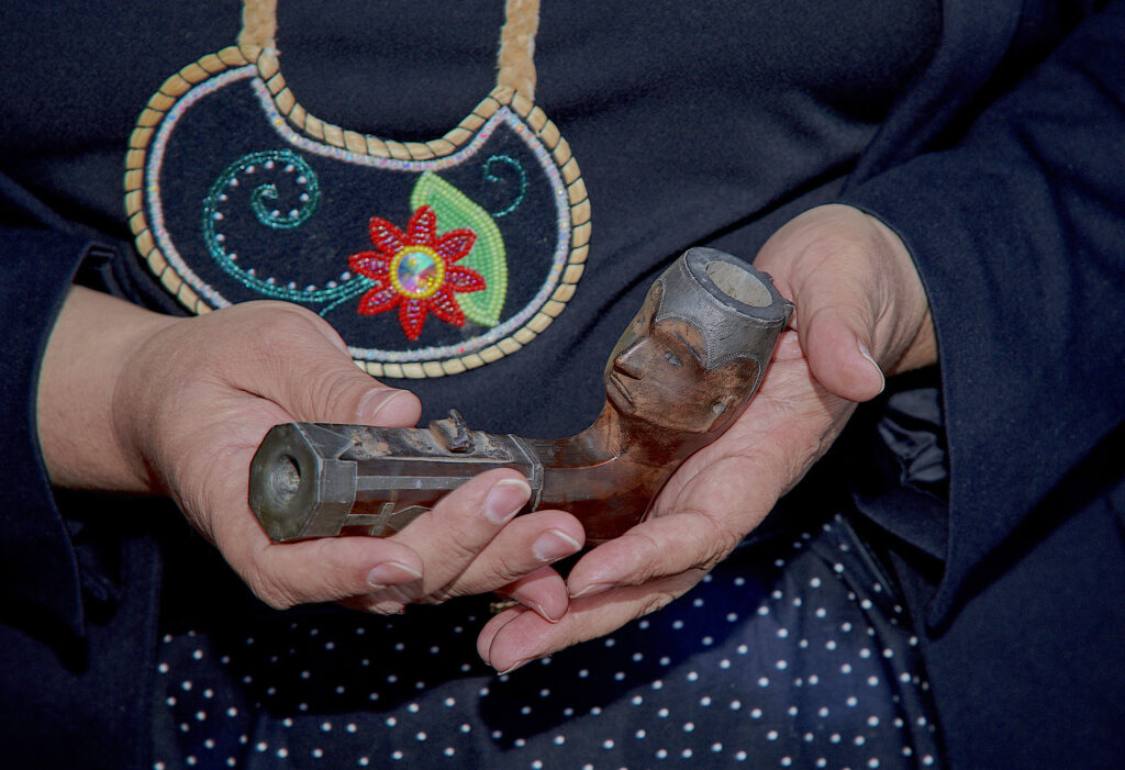 A close-up image features the hands of a person wearing a black top with a colorful beaded necklace cradling a wooden pipe with a face carved into its top end.